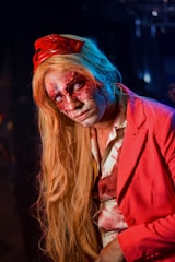 A person dressed in a striking, horror-themed costume with intricate face makeup resembling wounds. They wear a bright red jacket over a similarly stained shirt and have long blonde hair. A red hat or accessory is on their head, complementing the rest of the outfit.
