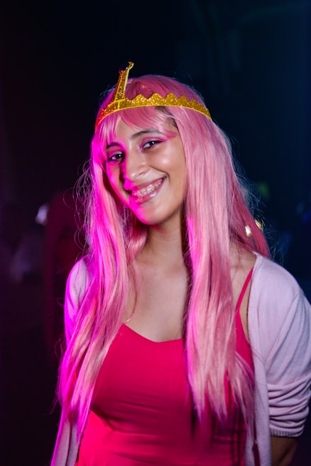 A person is wearing a pink wig and a yellow crown, smiling in a well-lit photo. The background is dark, highlighting the brightly colored costume. The individual is dressed in a pink outfit, enhancing the theme of the attire.