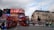 A busy urban scene with a large electronic billboard displaying advertisements and a historic building with classical architecture. Two red double-decker buses are prominent in the foreground, surrounded by a bustling crowd. The billboard shows various advertisements, including a prominent Coca-Cola ad. The sky is mostly cloudy with some patches of blue.