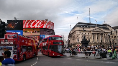 A busy urban scene with a large electronic billboard displaying advertisements and a historic building with classical architecture. Two red double-decker buses are prominent in the foreground, surrounded by a bustling crowd. The billboard shows various advertisements, including a prominent Coca-Cola ad. The sky is mostly cloudy with some patches of blue.