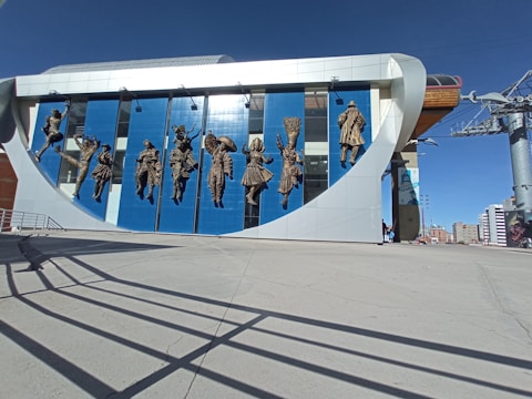 A modern building facade featuring bronze sculptures of people in traditional costumes, each posed dynamically against a backdrop of blue and white panels. The clear sky and architectural elements suggest an urban environment.