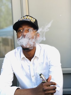 A man in a white shirt is exhaling vapor, creating a cloud of smoke in front of his face. He is wearing a black cap with lettering on it and holding a vaping device in his hand.