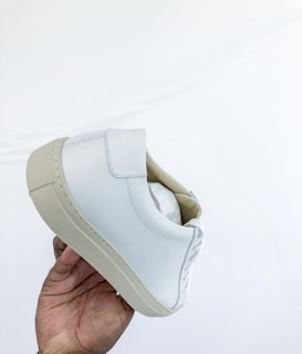 A hand is holding up a white leather sneaker with beige details, featuring clean stitching and a sleek design. The background is a simple white, emphasizing the minimalistic style of the shoe.