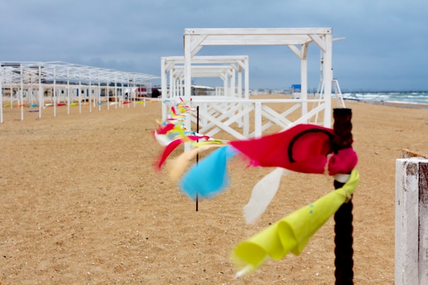 A beach scene with sandy shores and a series of white wooden structures, possibly beach cabanas or pergolas, lining the coast. Colorful ribbons are tied to a post in the foreground, fluttering in the wind, adding liveliness to the otherwise overcast setting. The ocean is visible in the background beneath a cloudy sky, suggesting a windy day.