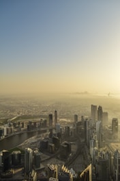 A sprawling urban cityscape featuring numerous modern skyscrapers stretching across the horizon under a hazy sky. The city is intersected by a winding river, and the buildings are densely packed, indicating a bustling metropolis.