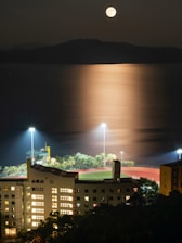 A brightly lit urban building stands in contrast to the dark night sky, with an illuminated sports field nearby. The full moon casts a shimmering reflection over the calm waters, creating a serene and tranquil atmosphere. Palm trees surround the sports field, which is bathed in artificial light from tall floodlights.
