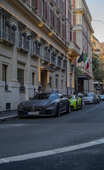 A street scene with luxury cars parked along the curb. The building features classic architecture with ornate balconies and a row of potted plants. Flags are hanging above the entrance, and the colors of the building are warm and inviting.