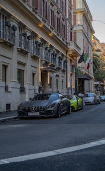 A street scene with luxury cars parked along the curb. The building features classic architecture with ornate balconies and a row of potted plants. Flags are hanging above the entrance, and the colors of the building are warm and inviting.