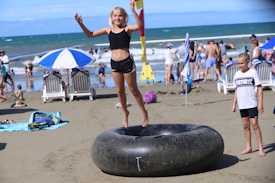 A child is joyfully jumping on a large rubber inner tube on a sandy beach. The beach is lively with people relaxing on lounge chairs under umbrellas and playing by the ocean. The background shows the sea waves hitting the shore under a clear blue sky.