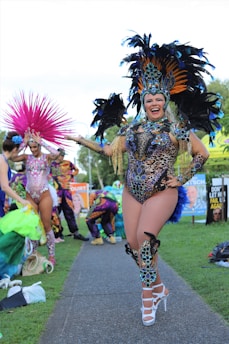 A person is walking along a path dressed in a vibrant and elaborate carnival costume with intricate beadwork and large feathered headgear. The costume features a leopard print design. In the background, other participants in colorful outfits are visible, preparing for an event.