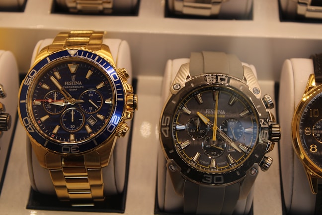 Two luxury watches are displayed in a well-lit watch case. The watch on the left features a gold band and case with a blue bezel and watch face, with multiple subdials and detailing. The watch on the right has a metallic look with a silver and grey appearance, including a grey band and several subdials.