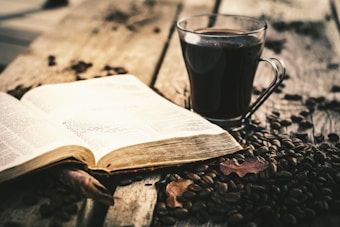 An open book lies on a rustic wooden table beside a glass mug filled with dark coffee. Coffee beans are scattered across the table, contributing to the cozy atmosphere. The weathered pages of the book and the wooden texture enhance the warm, inviting feel of the scene.