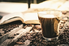 A clear glass mug filled with coffee is placed on a wooden surface scattered with coffee beans. In the background, an open book lies casually, suggesting a relaxed and cozy atmosphere.