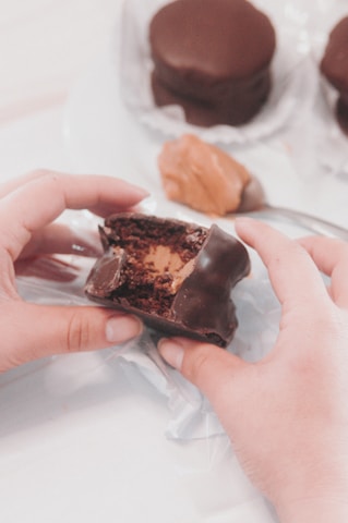 A close-up of hands holding a chocolate-covered snack with a creamy filling. The snack has been partially bitten, revealing its textured interior. In the background, there are other similar snacks and a dollop of peanut butter on a spoon.