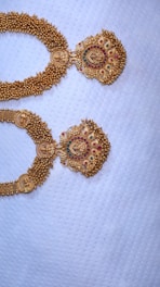 Two intricately designed gold necklaces with detailed patterns, featuring red and green gemstones, set against a textured white fabric background.