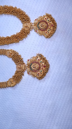 Two intricately designed gold necklaces with detailed patterns, featuring red and green gemstones, set against a textured white fabric background.