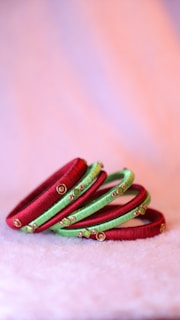 A set of colorful bangles, primarily red and green, are adorned with intricate embellishments and placed side by side on a plush, soft surface. The background features a gentle gradient of pink and purple tones, creating a soft and elegant appearance.
