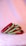 A set of colorful bangles, primarily red and green, are adorned with intricate embellishments and placed side by side on a plush, soft surface. The background features a gentle gradient of pink and purple tones, creating a soft and elegant appearance.