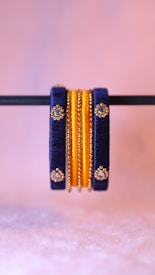 A pair of intricately designed bangles featuring dark blue and gold elements. The bangles are adorned with small, reflective gem-like decorations and are prominently displayed against a soft pink blurred background.