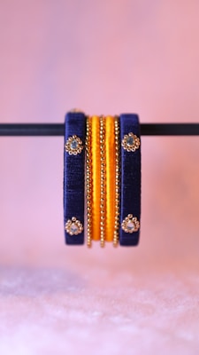 A pair of intricately designed bangles featuring dark blue and gold elements. The bangles are adorned with small, reflective gem-like decorations and are prominently displayed against a soft pink blurred background.