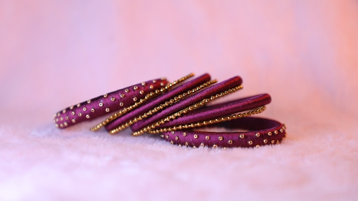 A set of purple and gold bangles placed on a soft, white surface. The bangles are adorned with intricate golden beads and patterns, creating a striking contrast against the purple. The background is soft and blurred, accentuating the details on the jewelry.