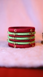 Several colorful bangles are neatly stacked on a soft, fuzzy surface. The bangles feature alternating shades of red and green with intricate gold embellishments and small embedded stones, creating a vibrant and traditional aesthetic.