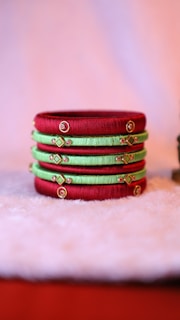 Several colorful bangles are neatly stacked on a soft, fuzzy surface. The bangles feature alternating shades of red and green with intricate gold embellishments and small embedded stones, creating a vibrant and traditional aesthetic.