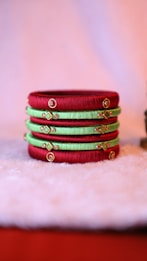 Several colorful bangles are neatly stacked on a soft, fuzzy surface. The bangles feature alternating shades of red and green with intricate gold embellishments and small embedded stones, creating a vibrant and traditional aesthetic.