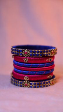 A stack of intricately designed bangles, featuring a combination of rich blue and deep red colors. The bangles are embellished with golden and pink square patterns, providing an elegant and ornate appearance against a soft, blurred background.