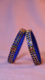 Two intricately designed blue bangles adorned with square gold and pink decorations are positioned upright against a softly blurred, warm-toned background.