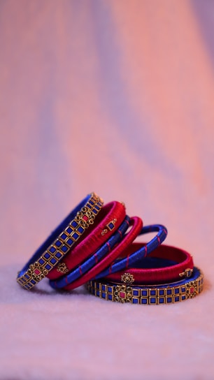 A set of stacked bangles in vibrant red and blue hues adorned with intricate gold detailing and embellishments. The bangles are arranged artistically on a soft, light-colored surface, creating a visually appealing contrast against the warm, blurred background.