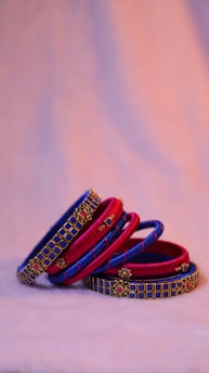 A set of stacked bangles in vibrant red and blue hues adorned with intricate gold detailing and embellishments. The bangles are arranged artistically on a soft, light-colored surface, creating a visually appealing contrast against the warm, blurred background.