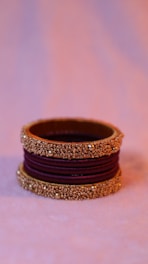 A decorative bangle with intricate gold beadwork and a purple fabric center. The bangle is elegantly crafted with three sections of gold embellishments separated by deep purple fabric. The background is softly blurred, highlighting the bangle.