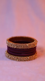 A decorative bangle with intricate gold beadwork and a purple fabric center. The bangle is elegantly crafted with three sections of gold embellishments separated by deep purple fabric. The background is softly blurred, highlighting the bangle.