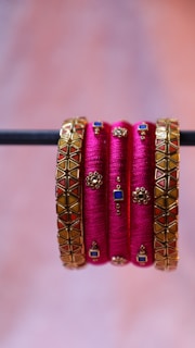 A set of vibrant and decorative pink bangles, embellished with intricate golden designs featuring blue and red geometric patterns, are elegantly displayed hanging from a horizontal rod.