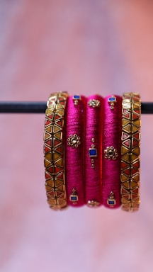 A set of vibrant and decorative pink bangles, embellished with intricate golden designs featuring blue and red geometric patterns, are elegantly displayed hanging from a horizontal rod.