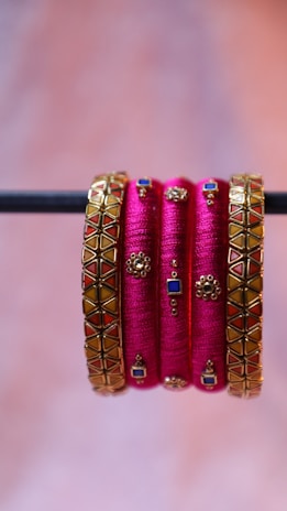 A set of vibrant and decorative pink bangles, embellished with intricate golden designs featuring blue and red geometric patterns, are elegantly displayed hanging from a horizontal rod.
