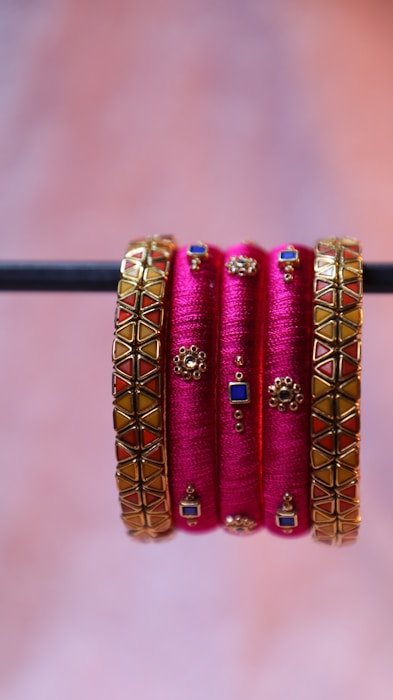 A set of vibrant and decorative pink bangles, embellished with intricate golden designs featuring blue and red geometric patterns, are elegantly displayed hanging from a horizontal rod.