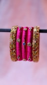 A set of vibrant pink bangles with intricate gold embellishments and geometric designs. Some bangles have squares of blue and red set into the design and small, detailed gold motifs throughout.