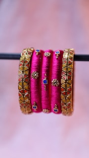 A set of vibrant pink bangles with intricate gold embellishments and geometric designs. Some bangles have squares of blue and red set into the design and small, detailed gold motifs throughout.