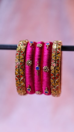 A set of vibrant pink bangles with intricate gold embellishments and geometric designs. Some bangles have squares of blue and red set into the design and small, detailed gold motifs throughout.