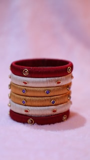 A decorative bangle crafted from red, gold, and beige threads, adorned with small colorful gems and gold accents, resting on a soft pastel background.