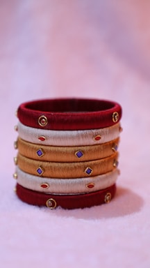 A decorative bangle crafted from red, gold, and beige threads, adorned with small colorful gems and gold accents, resting on a soft pastel background.
