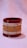 A decorative bangle crafted from red, gold, and beige threads, adorned with small colorful gems and gold accents, resting on a soft pastel background.
