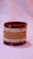 A decorative bangle crafted from red, gold, and beige threads, adorned with small colorful gems and gold accents, resting on a soft pastel background.