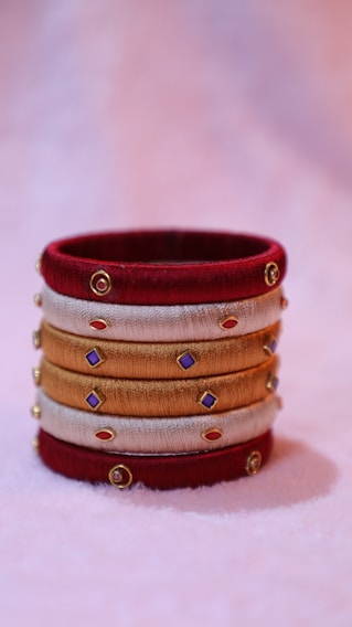 A decorative bangle crafted from red, gold, and beige threads, adorned with small colorful gems and gold accents, resting on a soft pastel background.