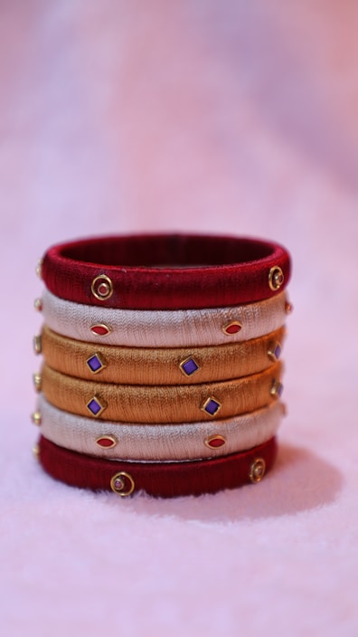 A decorative bangle crafted from red, gold, and beige threads, adorned with small colorful gems and gold accents, resting on a soft pastel background.