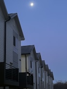 A row of modern, multi-story townhouses with white siding and dark balconies are aligned closely together. The sky above is a deep blue, with the moon visible and illuminating the scene.