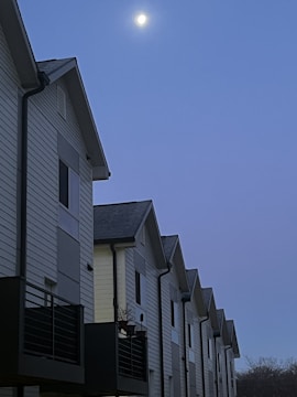 A row of modern, multi-story townhouses with white siding and dark balconies are aligned closely together. The sky above is a deep blue, with the moon visible and illuminating the scene.
