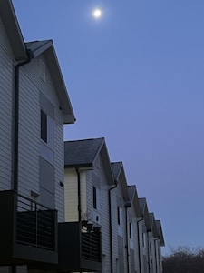A row of modern, multi-story townhouses with white siding and dark balconies are aligned closely together. The sky above is a deep blue, with the moon visible and illuminating the scene.